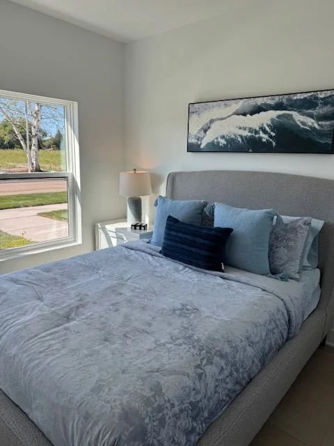 Bedroom with a gray bed, blue pillows, a lamp on a white nightstand, and a window showing a tree and sidewalk outside.
