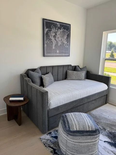 A gray upholstered daybed with dark gray and light gray pillows, situated in a bright room next to a window. A small wooden side table with books is beside it, and a round, woven ottoman in front. There is a black-framed map on the wall above the day