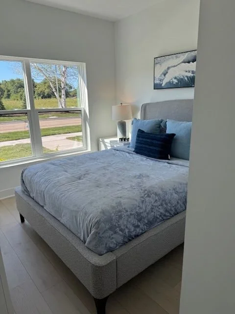A neat bedroom with a large bed, blue and navy pillows, a window showing trees outside, a nightstand with a lamp, and a framed abstract art piece on the wall.
