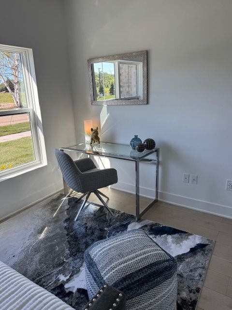 A small area with a desk by a window, a gray chair, and a mirror on the wall. The desk has decorative vases and a small lamp with a cat figure. There is a rug on the floor and a textured ottoman in the foreground.