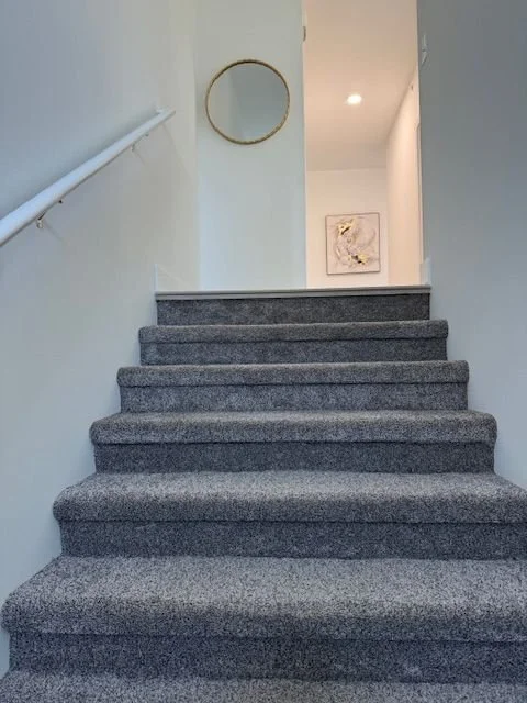Carpeted staircase leading to an upper level, with a white wall on the left and a light-colored wall on the right. There is a circular mirror on the wall at the top of the stairs and a framed picture in the background.