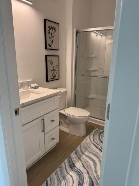 A small bathroom with white cabinetry, a toilet, a glass shower with built-in shelves, and framed botanical art on the wall.