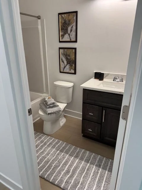 A small bathroom with a toilet, a dark wood vanity with a white countertop and sink, two black containers on the counter, a striped rug, and two framed botanical pictures on the wall.