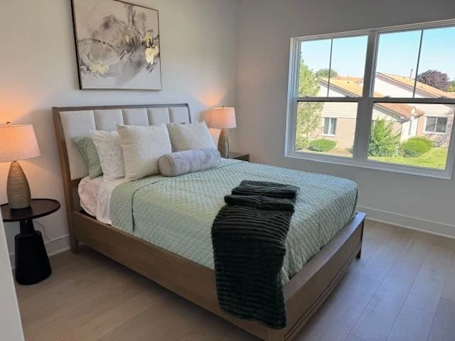 A neatly made king-sized bed with a light-colored quilt, multiple pillows, and a rolled bolster pillow. The bed is flanked by matching bedside tables with lamps. There is a large abstract painting above the headboard, and a window showing a suburban 