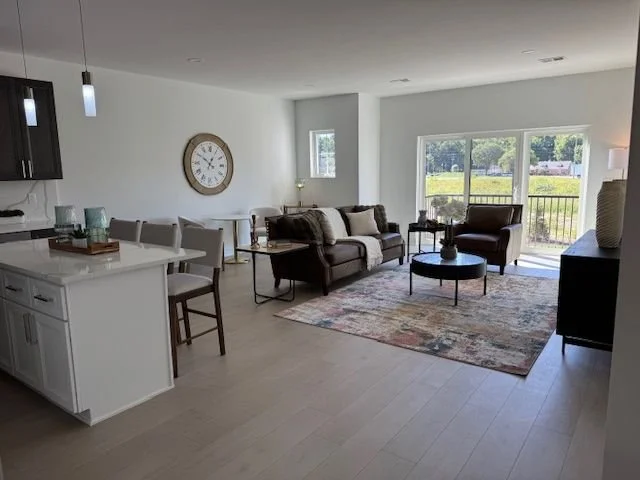 Living room with brown sofa, armchair, glass coffee table, rug, and large window overlooking greenery.