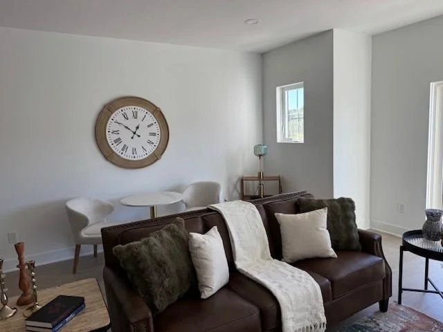 Living room with a brown leather sofa, white pillows, a cream throw blanket, wall clock, small window, side table with lamp, and decorative candles.