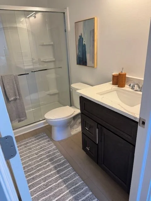 Bathroom with glass shower enclosure, toilet, dark vanity with marble countertop, and abstract art on the wall