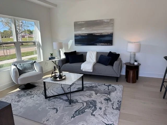 Living room with a gray sofa, black cushions, a white blanket, a marble-top coffee table, a patterned area rug, a gray armchair, two white table lamps, a large abstract painting, and a window showing an outdoor scene.