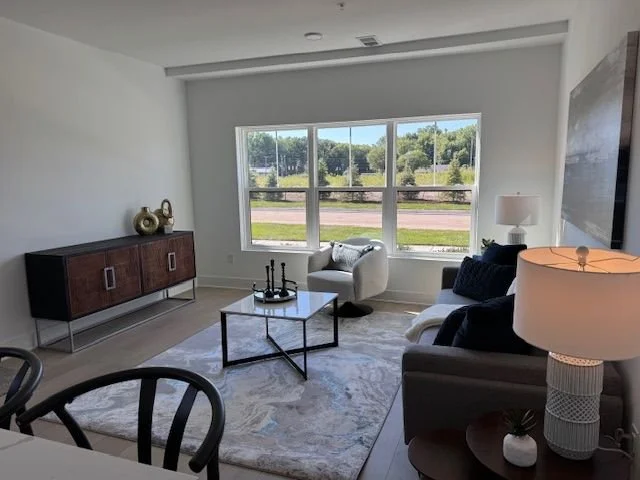 Living room with large window, white walls, gray sofa with dark blue pillows, white armchair, black coffee table, side table with white lamp, and a dark sideboard with decorative items.