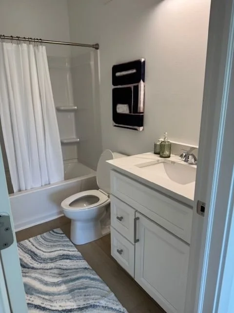 Bathroom with a bathtub and shower, toilet, white vanity with sink, mirror, and black wall art, on a wood floor with a striped rug.