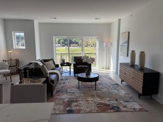 Living room with large windows, sofa, armchair, coffee table, side table with flowers, floor lamp, colorful area rug, and decorative vases on a sideboard.
