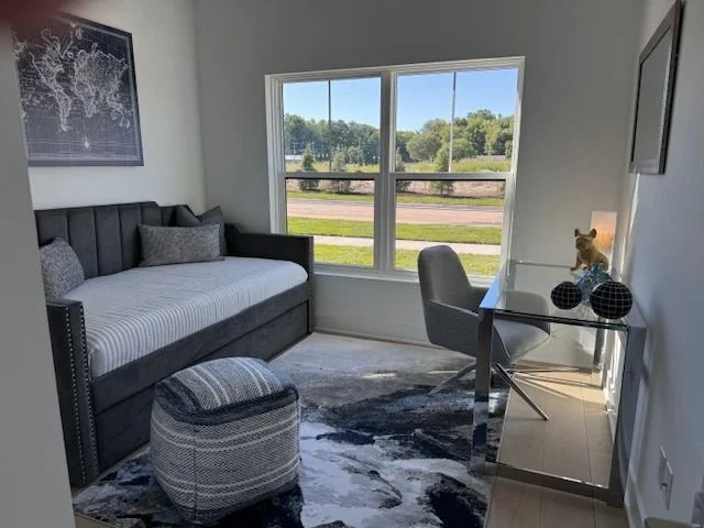 A cozy room with a black daybed, a striped ottoman, and a glass desk near a large window with a view of a grassy area and trees outside. The desk has decorative objects and a small stuffed animal.