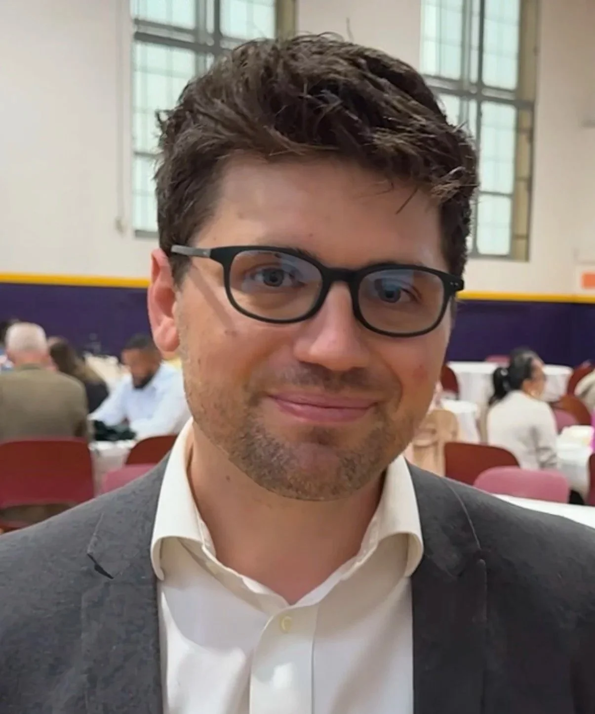 A man with dark brown hair, glasses, and a light beard smiling in a large indoor setting with tables and people seated.