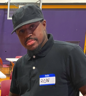 Young man wearing a black cap and black shirt with a name tag that says 'Hello RON' in a purple and orange indoor setting.