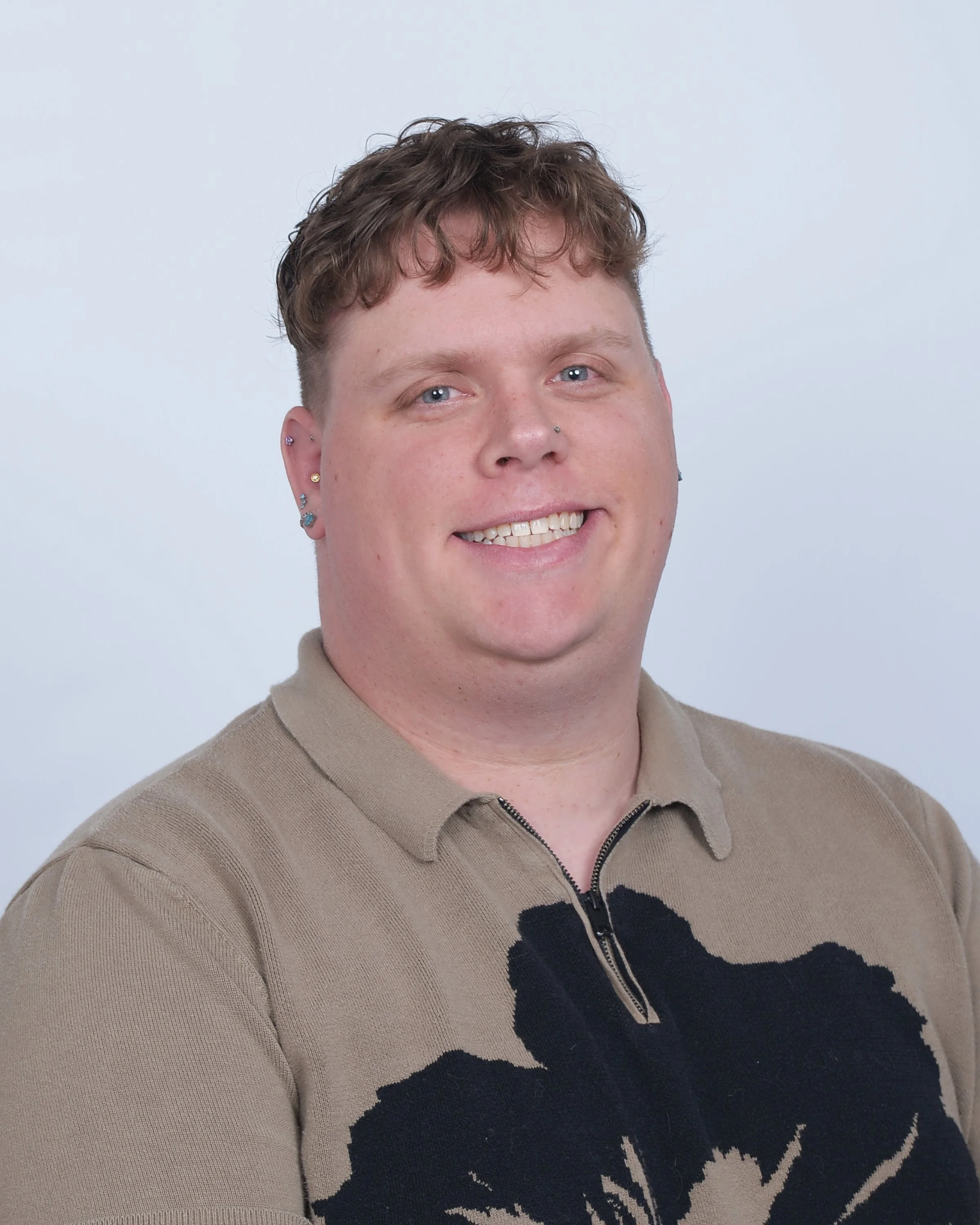 A young man smiling, wearing a beige polo shirt with a black graphic design, earrings, and a nose piercing against a plain light background.