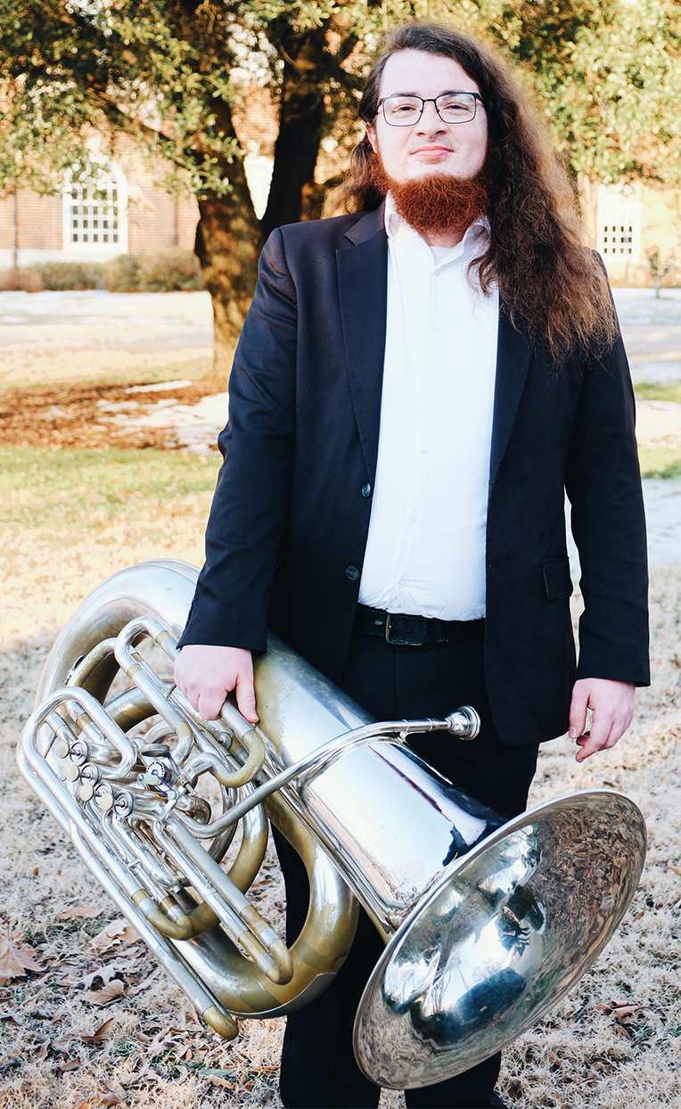 Man with long hair and a brown-red beard wearing glasses, a white shirt, and a dark blazer standing outdoors on a winter day holding a large silver tuba.