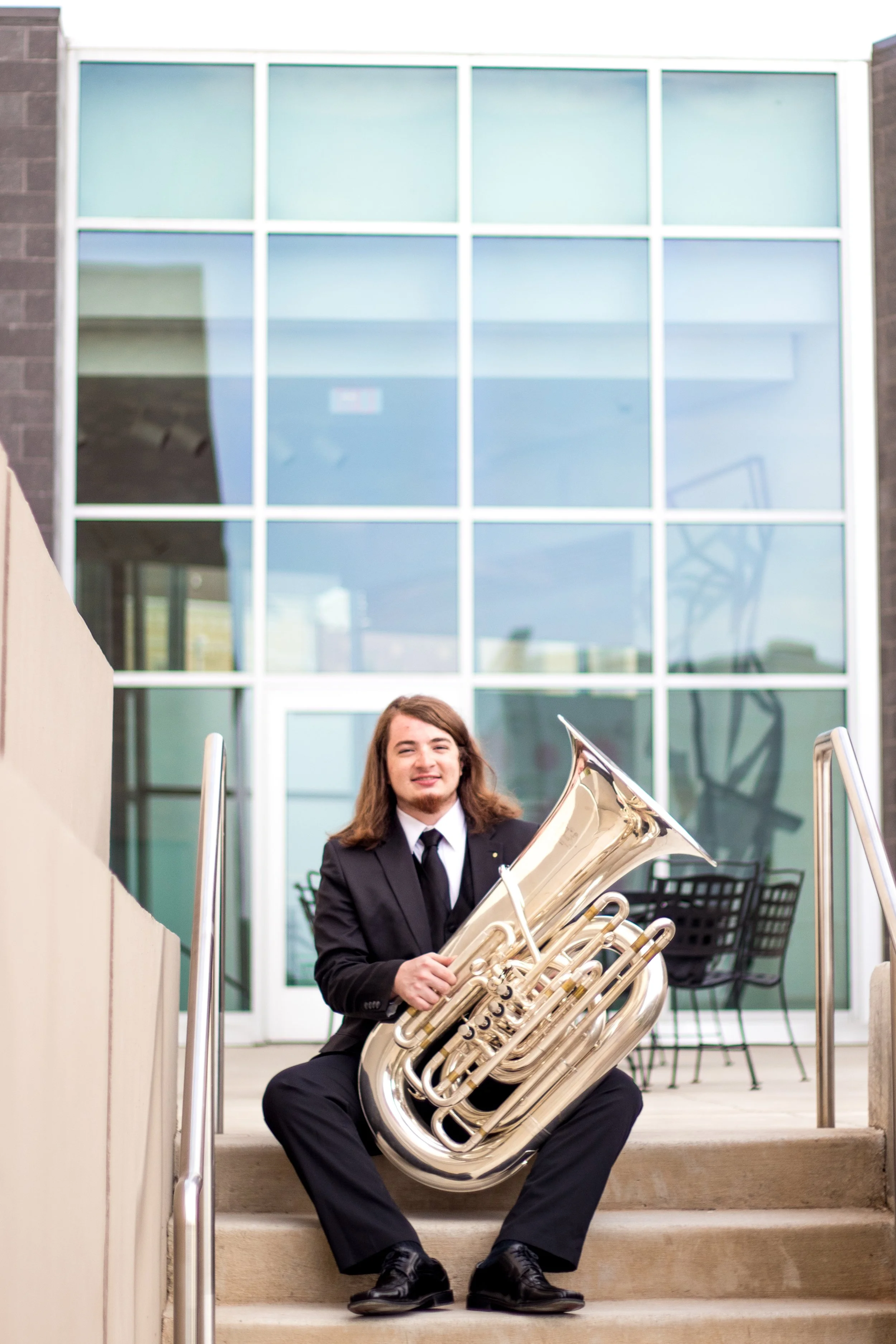 A young man in a black suit sitting on outdoor stairs, holding a large silver tuba, with a modern building with glass windows behind him.