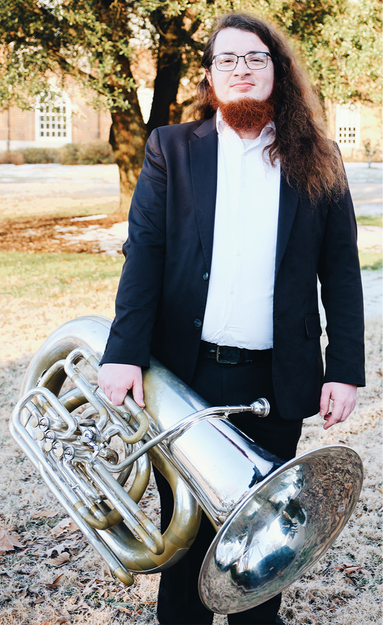 A man with long, curly brown hair and a full red beard holding a large brass tuba outside on a sunny day, standing in front of a tree and a building with windows.