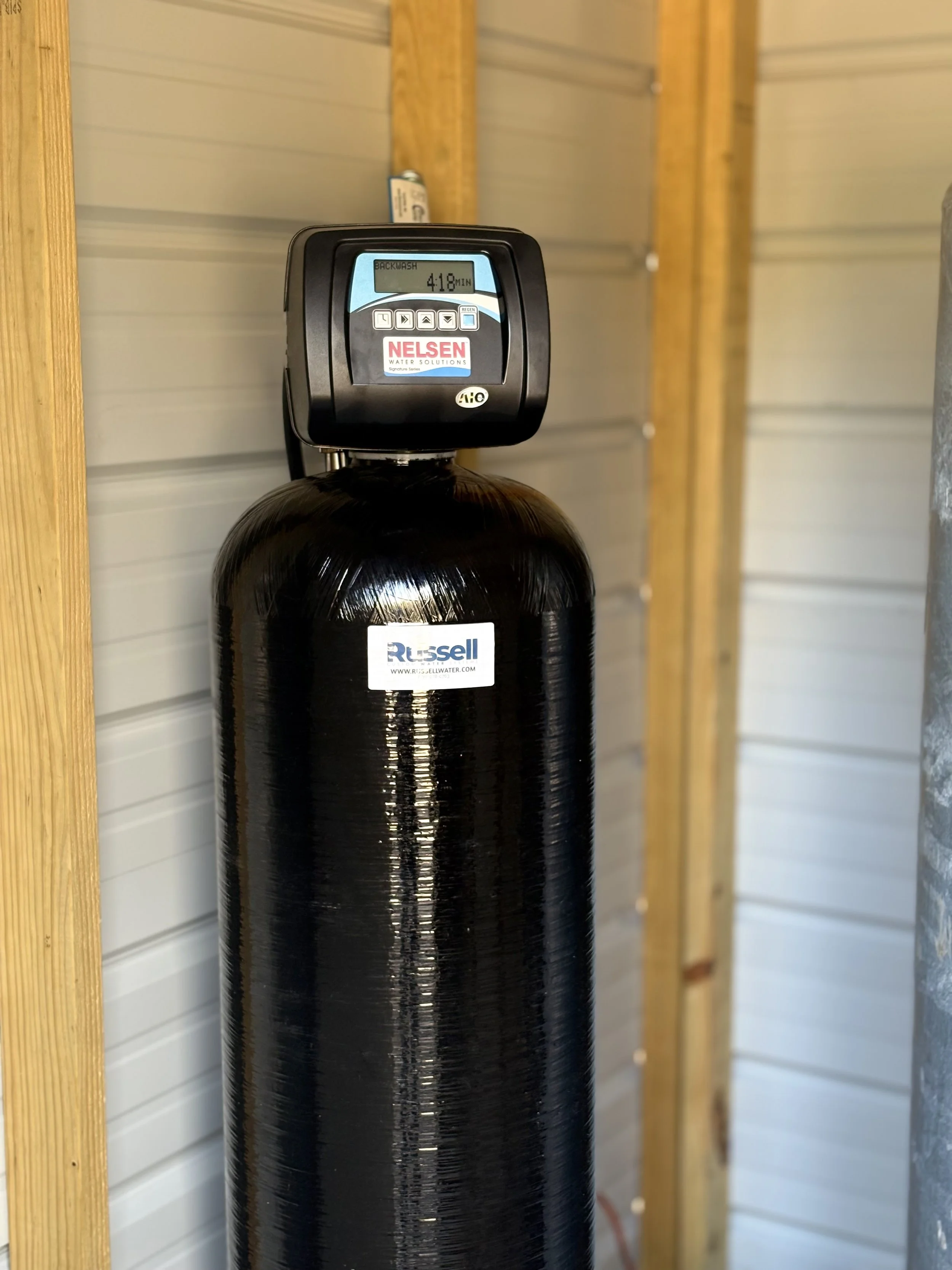 A black water filtration tank with a digital control head on top, installed in a wooden frame, with labels reading 'Russell' and 'NELSON'