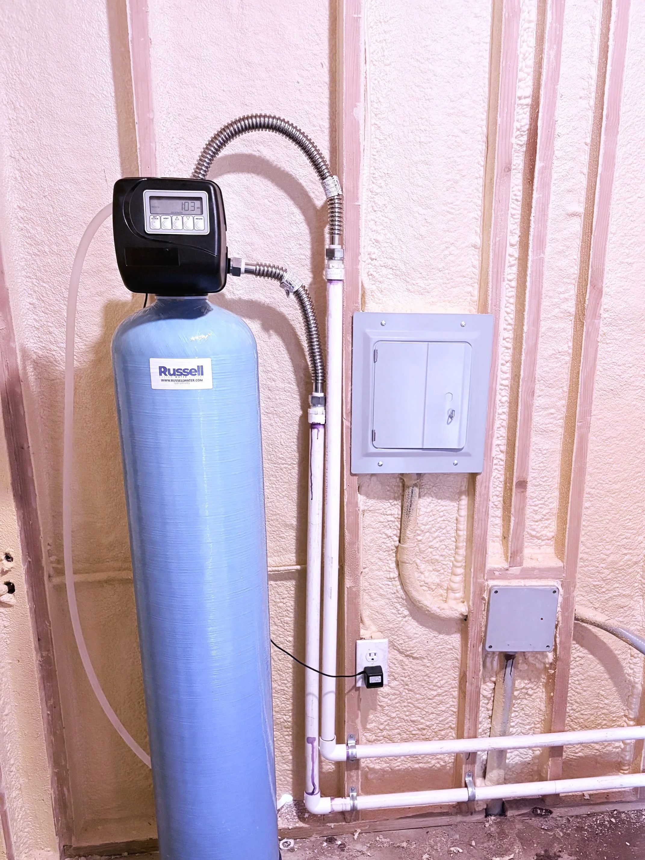 A water softener system in a utility room with pipes, control head, and electrical panel mounted on a wall.