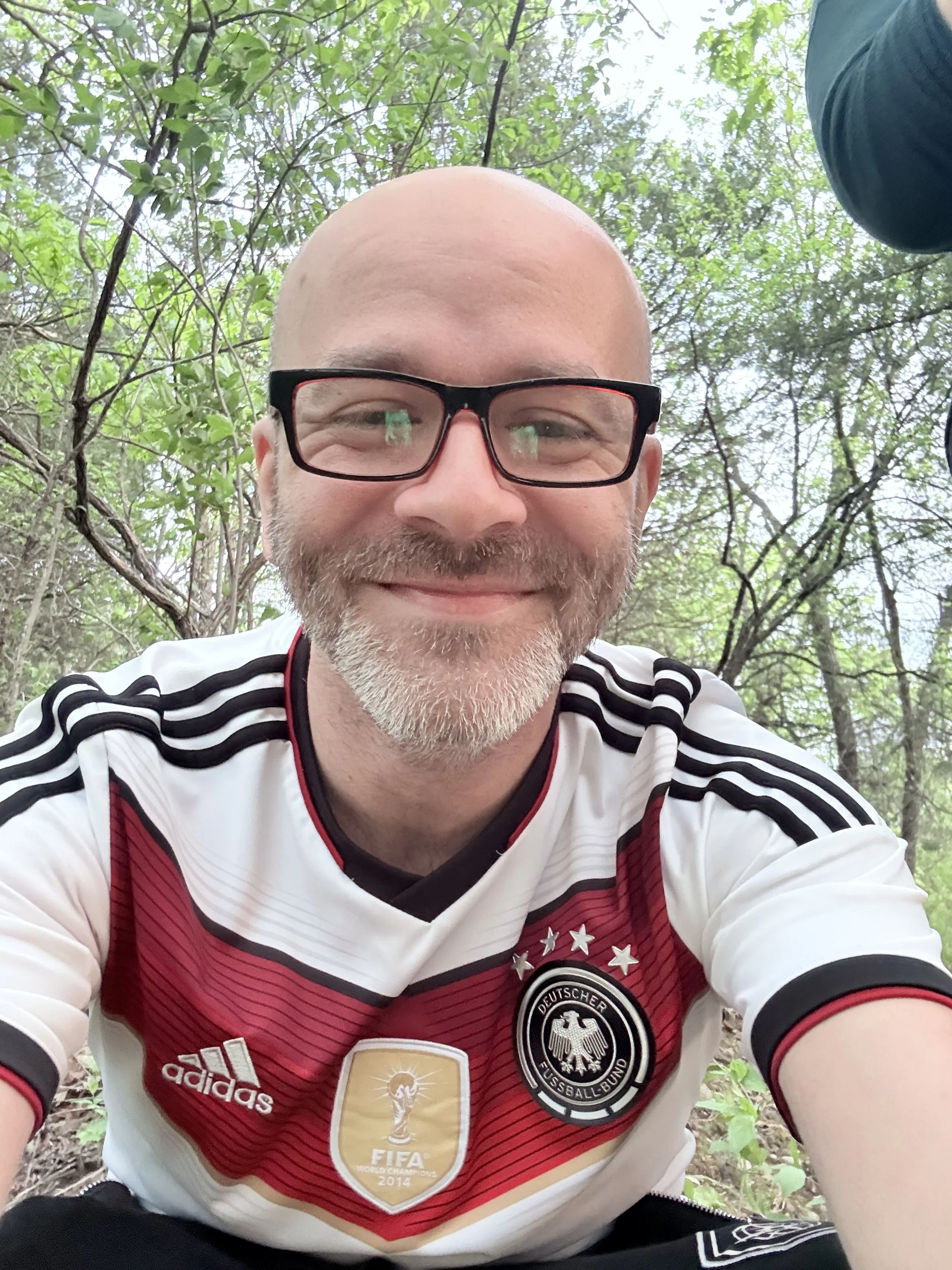 A man wearing glasses and a German national soccer team jersey taking a selfie outdoors among green trees.
