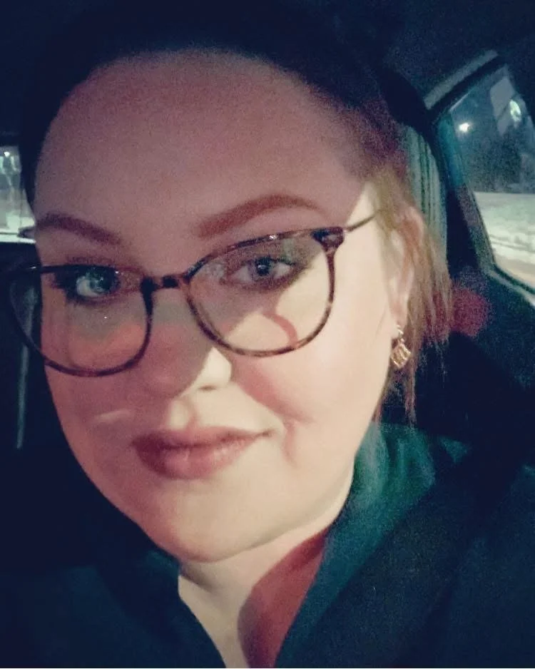 A woman with glasses, brown hair, and earrings sitting in a car at night.