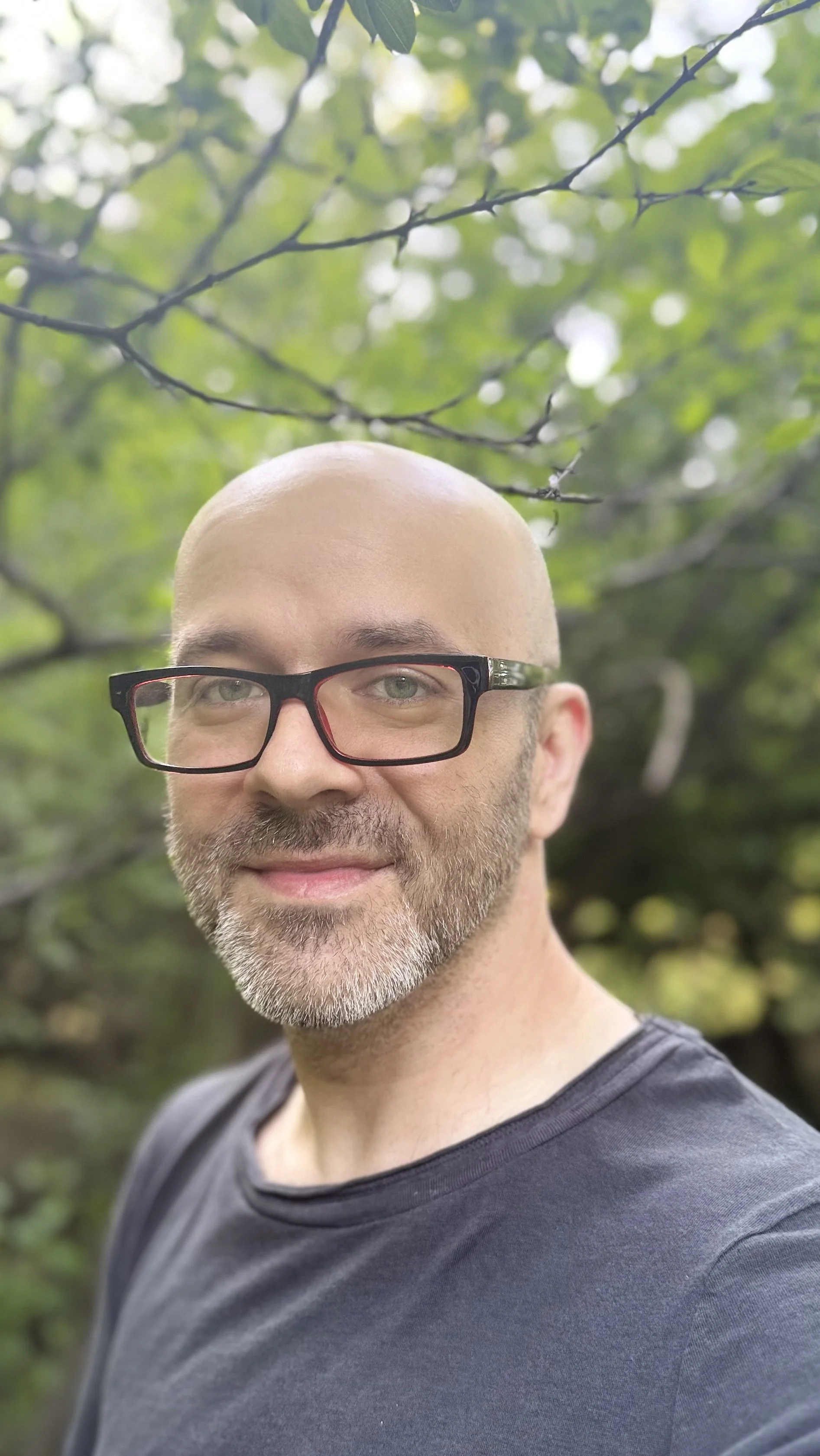 A man with a beard and glasses smiling outdoors, with green foliage and tree branches in the background.