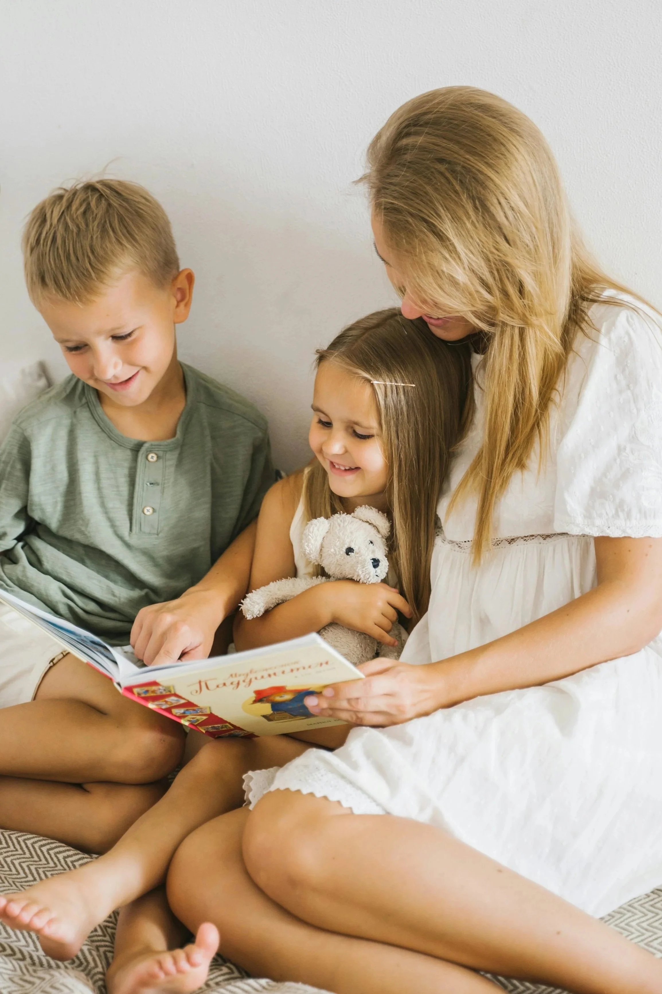 A woman and two children reading a book together, with the girl holding a stuffed animal, sitting on a bed against a plain white wall.