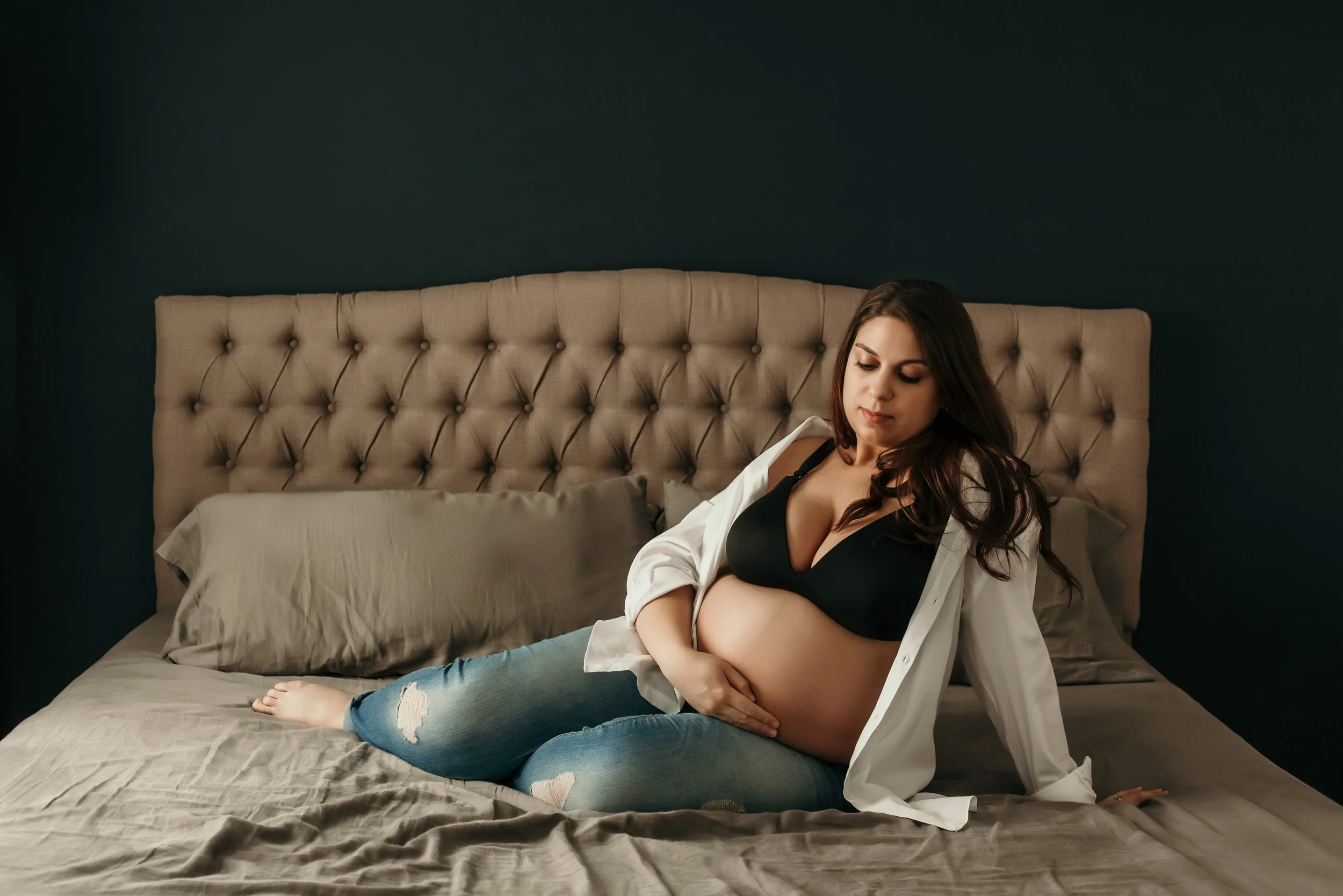 Pregnant woman sitting on bed, wearing a black bra, open white shirt, and ripped jeans, cradling her belly.