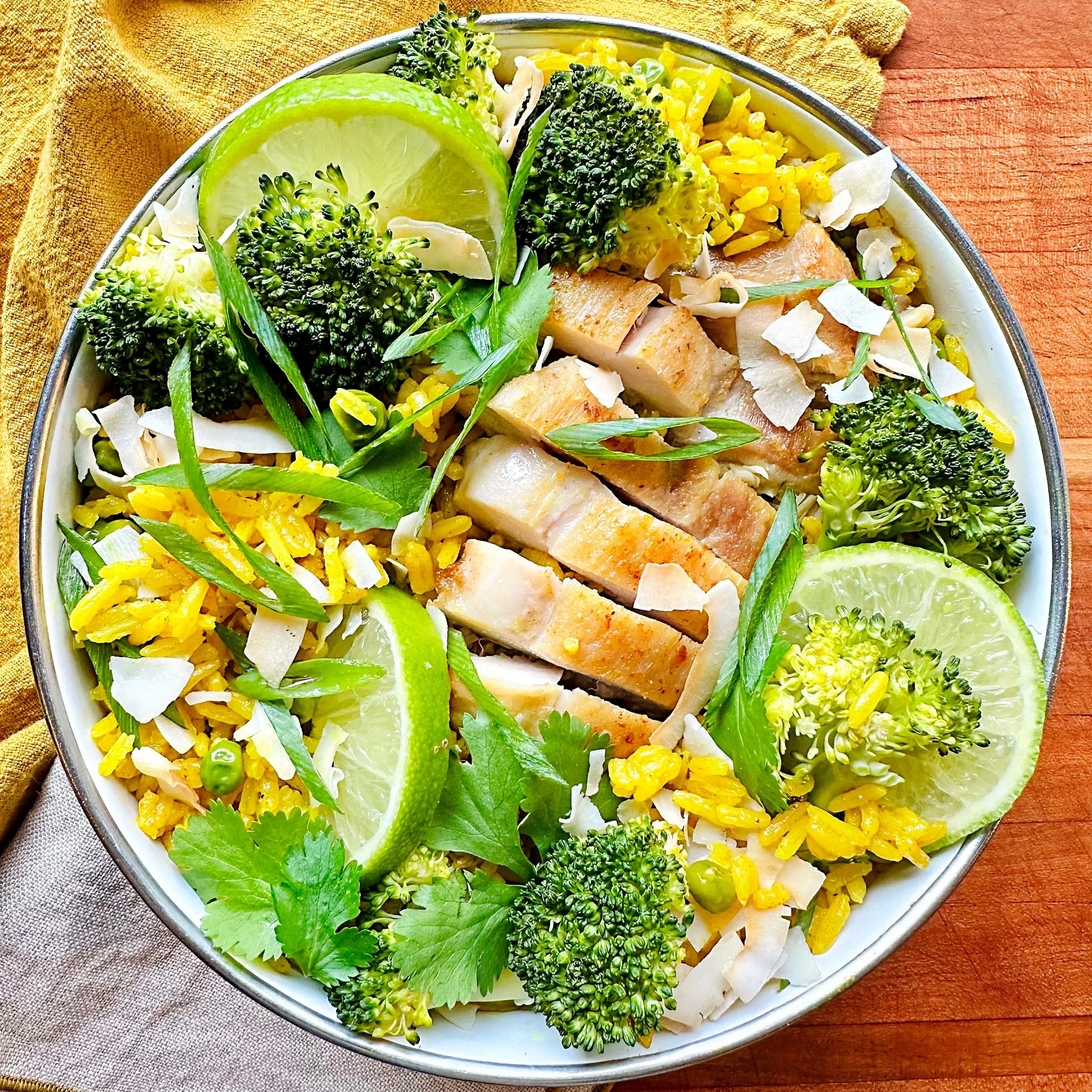 Salad bowl with grilled chicken, broccoli, lime wedges, rice, and fresh herbs on a wooden table.