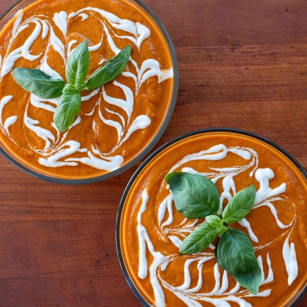 Two bowls of tomato soup garnished with basil leaves and drizzled with cream on top, placed on a wooden surface.