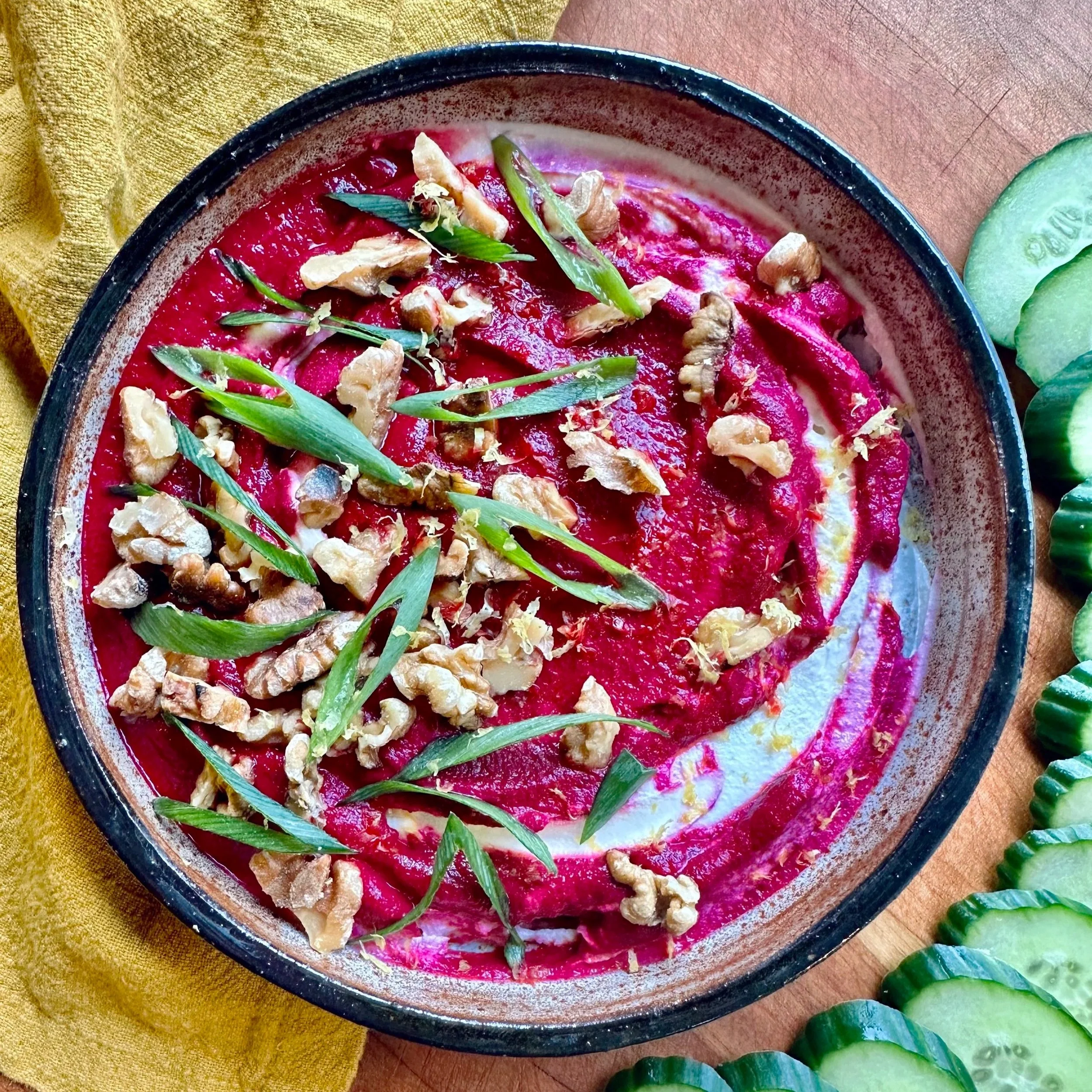 A bowl of beetroot hummus garnished with chopped nuts and fresh herbs, served alongside sliced cucumbers.