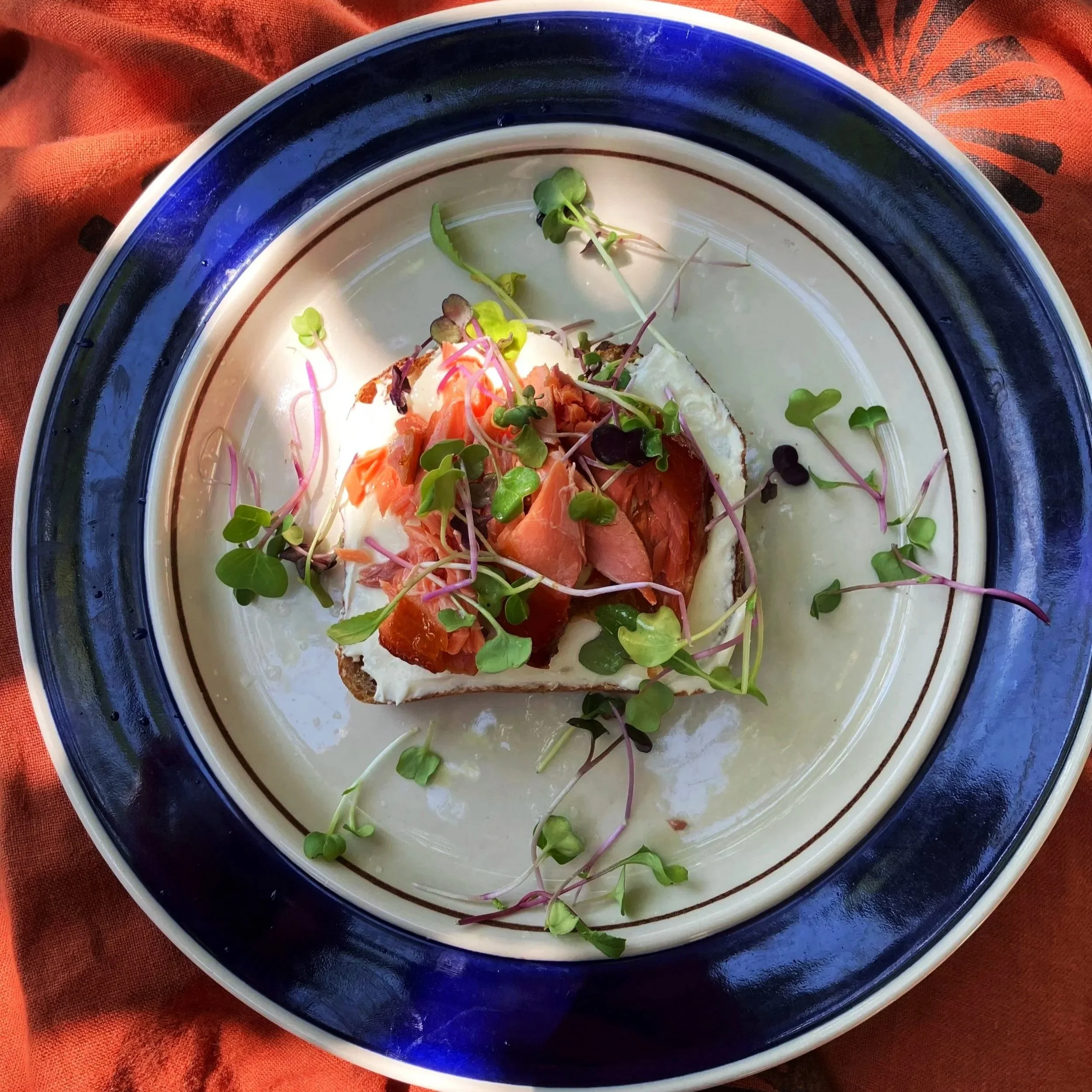 A plate with a slice of bread topped with cream cheese, smoked salmon, and microgreens, served on a decorative plate with a blue rim, on a reddish-orange cloth.