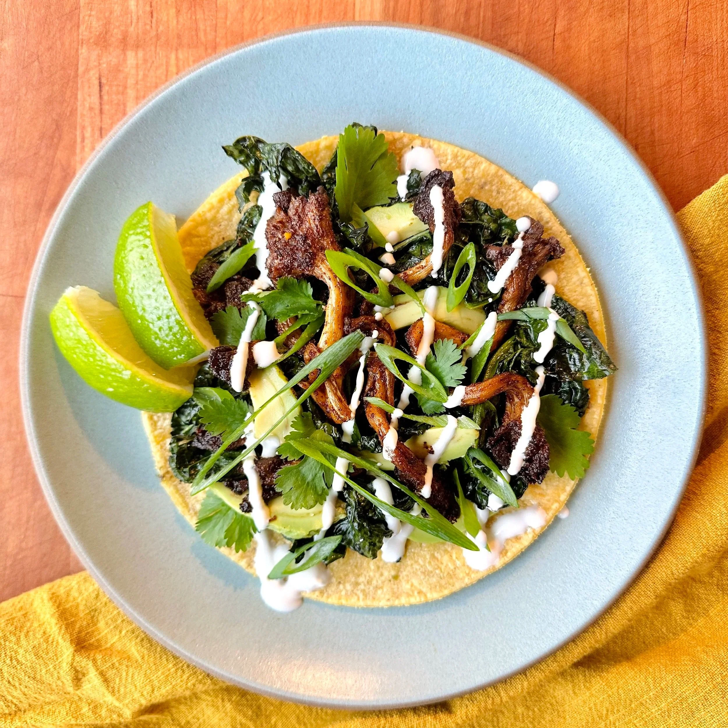 A plate of tacos topped with grilled meat, leafy greens, chopped herbs, drizzled with white sauce, with lime wedges on the side, on a yellow cloth with a wooden surface background.