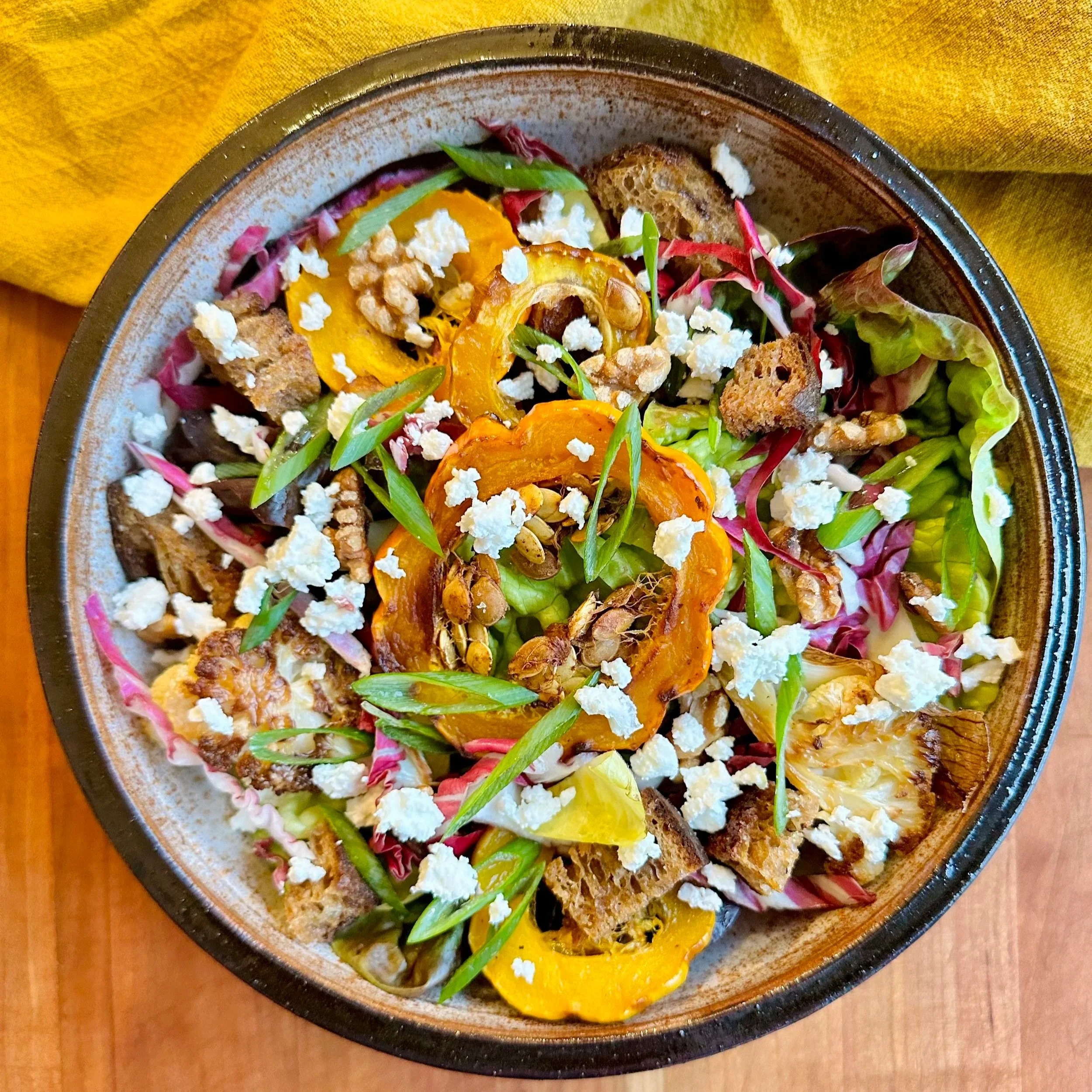 Colorful salad with roasted yellow and orange peppers, leafy greens, crumbled cheese, chopped nuts or seeds, bread crumbles, and thinly sliced green onions in a brown bowl on a wooden table.