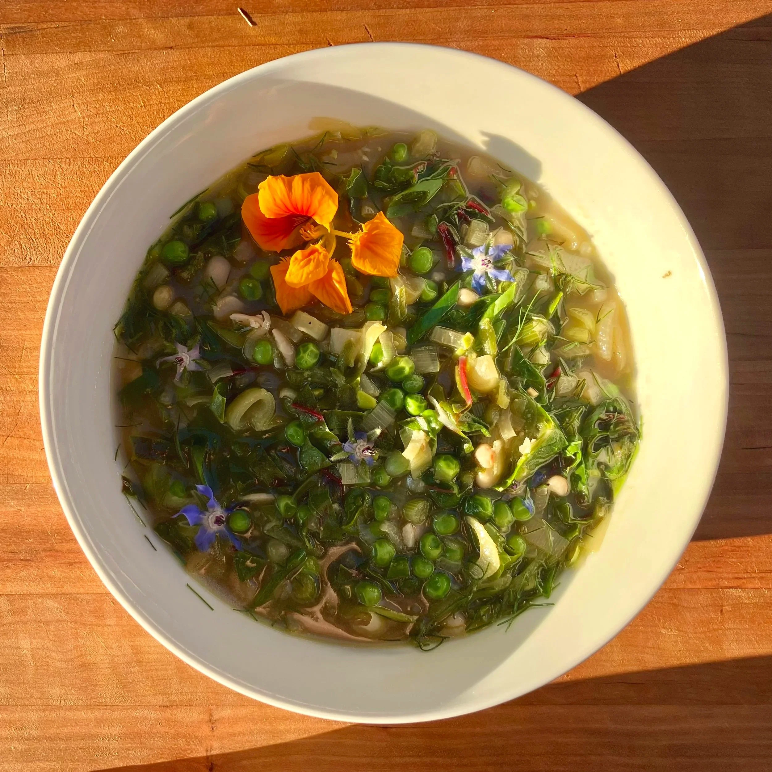 A bowl of vegetable soup garnished with edible flowers on a wooden surface.