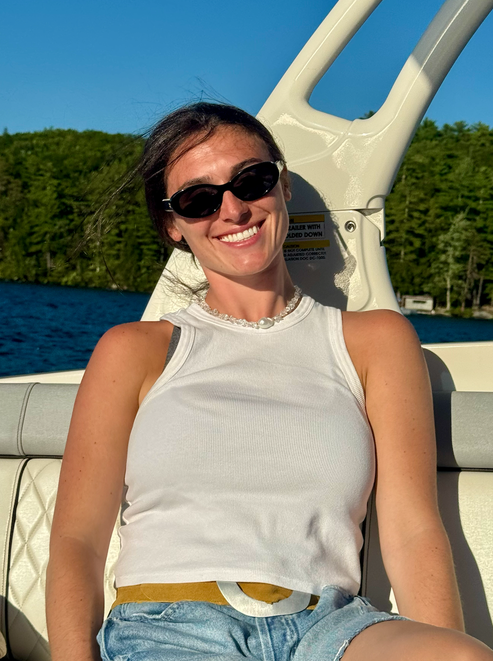 Smiling woman wearing sunglasses, tank top, and shorts sitting on a boat with water and trees in the background.