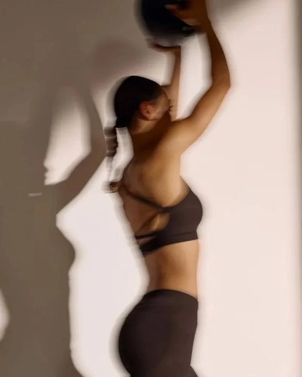 A woman with dark hair in a ponytail, wearing black workout clothing, is stretching or reaching up with both arms while standing against a wall. The photo is blurry and has artistic shadow effects.