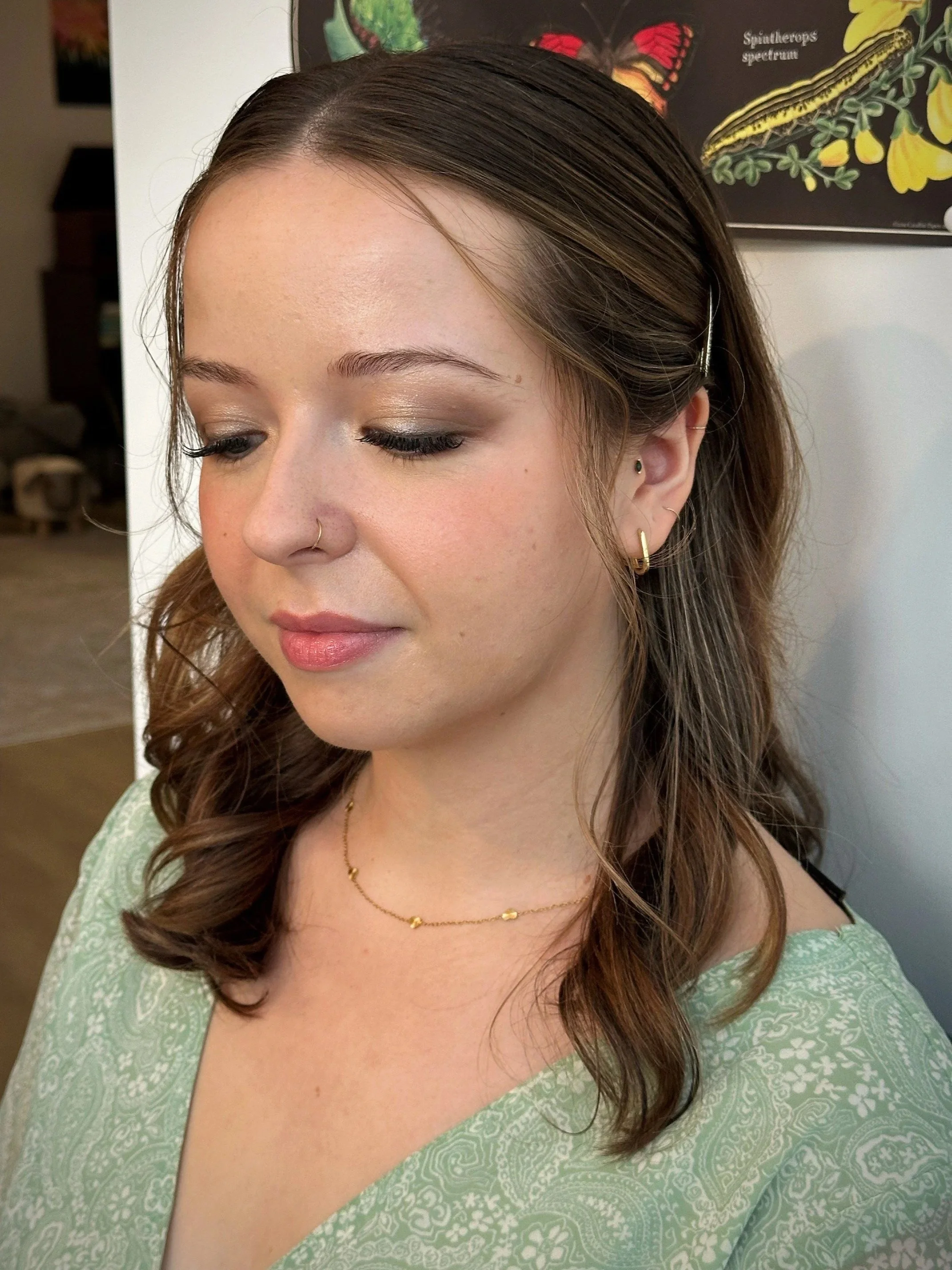 A woman with wavy brown hair, wearing makeup, a nose ring, gold earrings, a delicate gold necklace, and a green patterned top, is looking down.