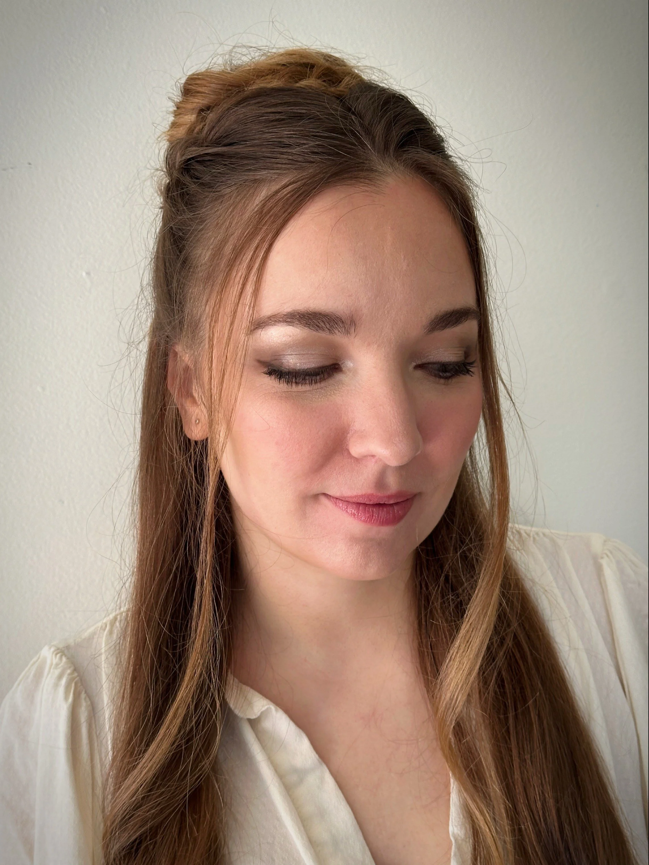 A woman with long, light brown hair styled in loose waves and a top bun, wearing makeup with eyeshadow and lipstick, looking down against a plain white wall background.