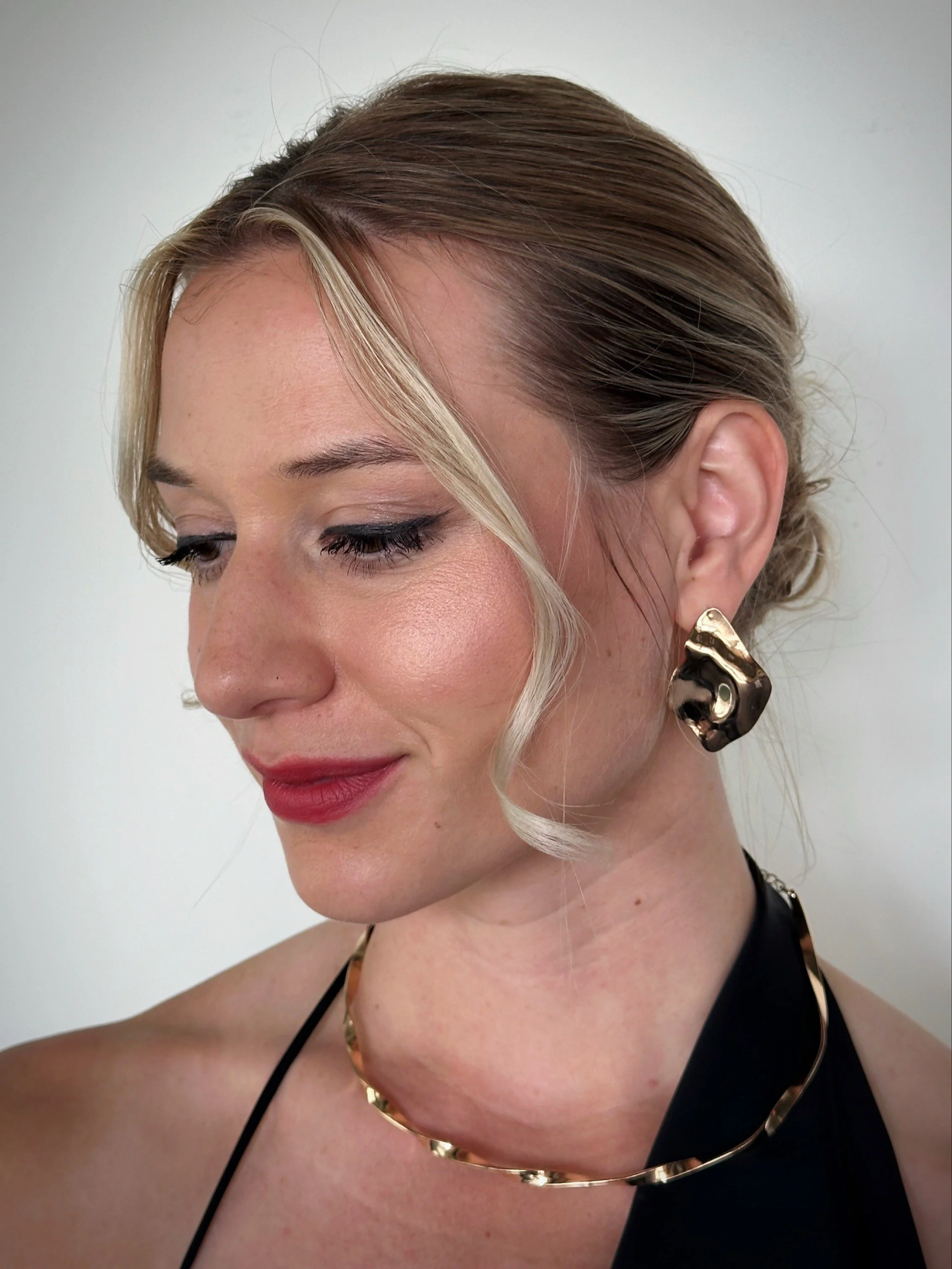 Close-up of a woman with blonde hair styled in an updo, wearing bold earrings and a black strap dress, with makeup including red lipstick and winged eyeliner, against a neutral background.