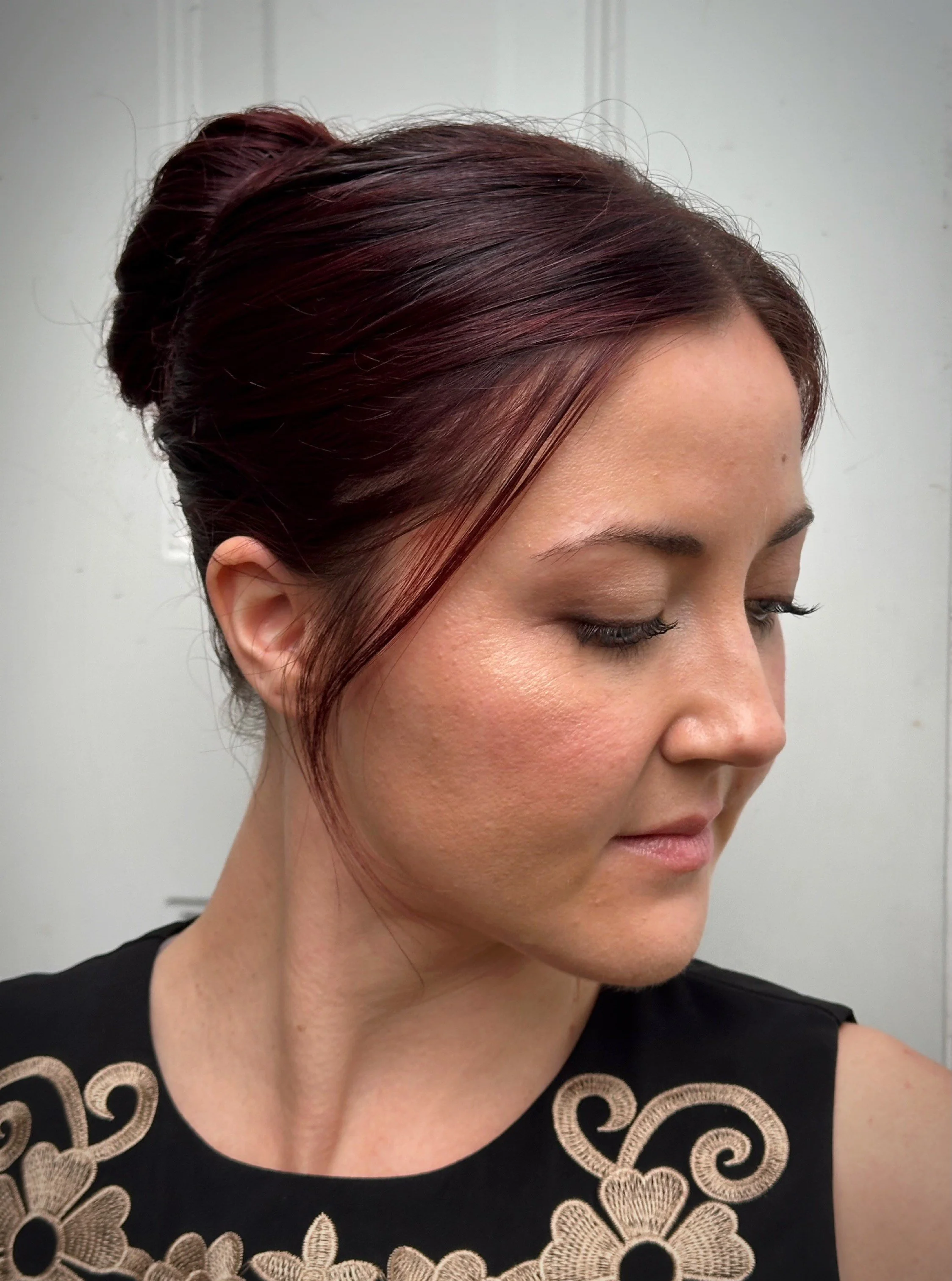A woman with burgundy hair styled in a low updo, wearing a black top with gold embroidery, looking down and to the side against a plain background.