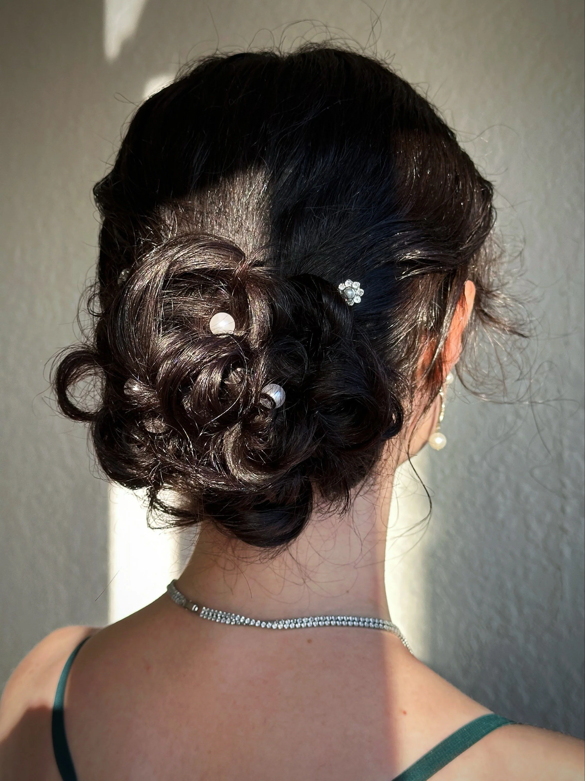Back view of a woman with an elegant updo hairstyle decorated with pearl and rhinestone hair accessories, wearing a diamond necklace, pearl earrings, and a green strap dress.