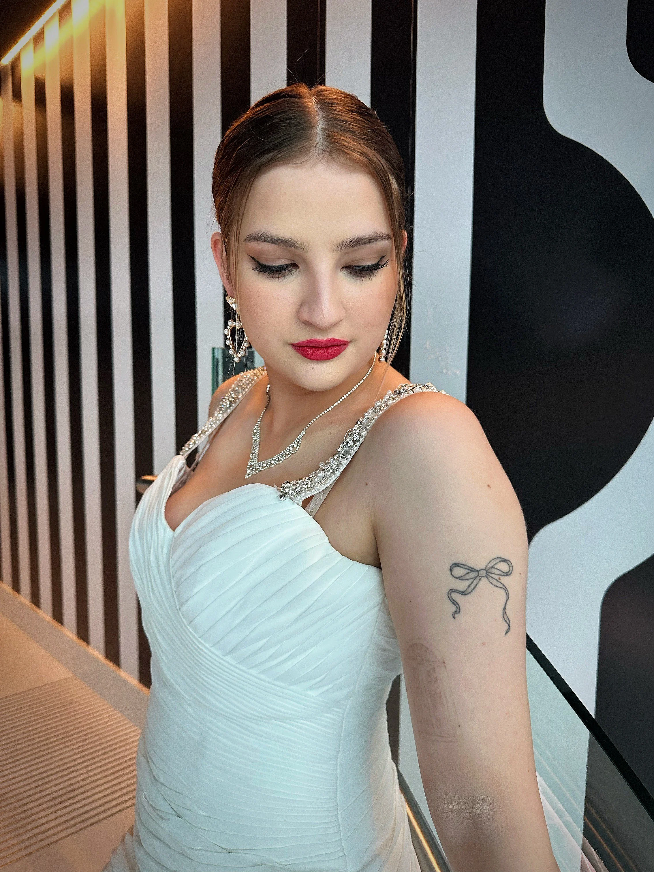 A young woman in a white dress with jewelry, looking down, standing indoors with a black and white striped wall behind her.