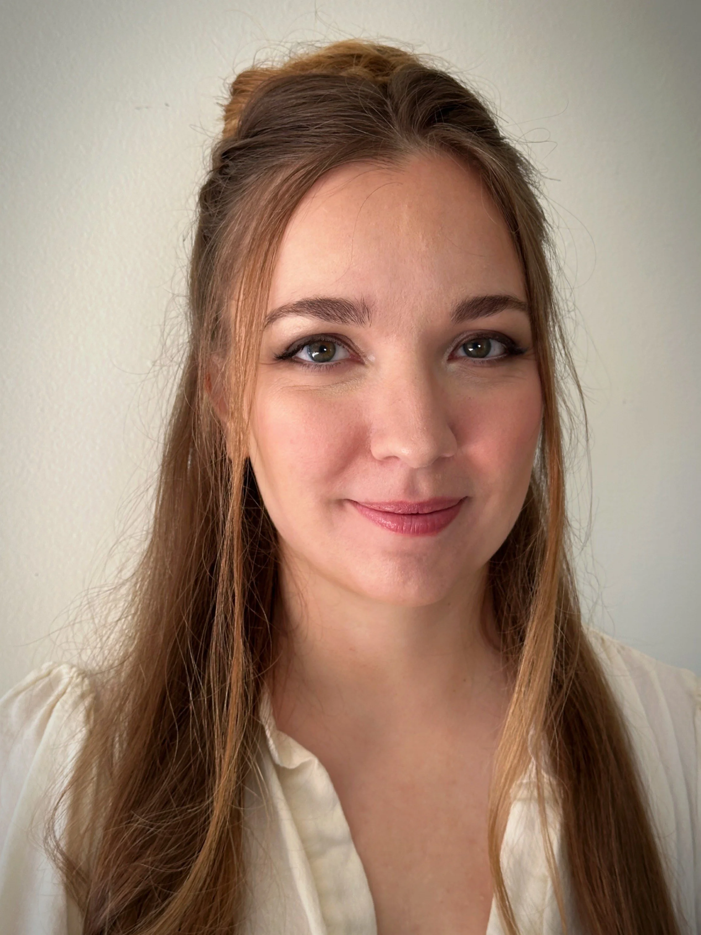 A young woman with long light brown hair, styled half-up, smiling at the camera against a plain light-colored wall.