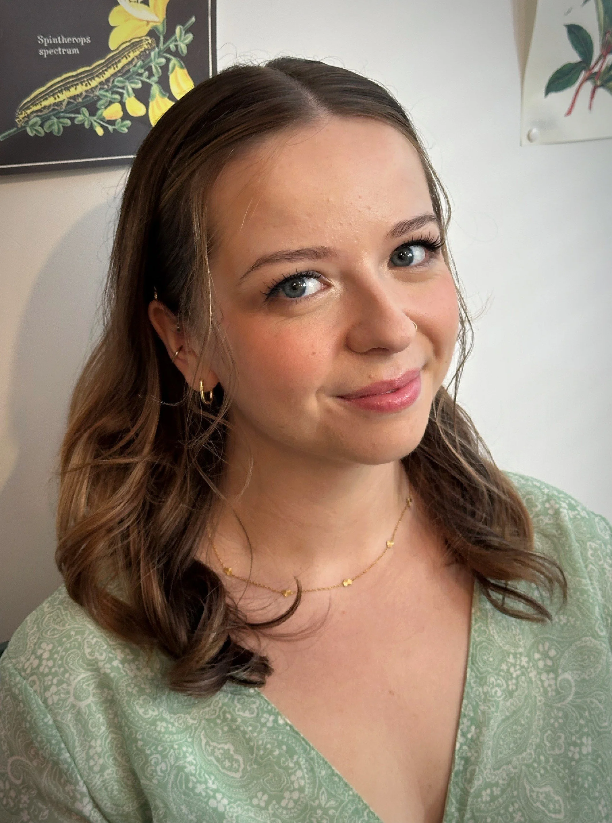 A young woman with wavy brown hair and blue eyes, wearing gold hoop earrings and a delicate gold necklace with small hearts. She is smiling slightly and is dressed in a light green patterned top. The background includes a black poster of a creature w