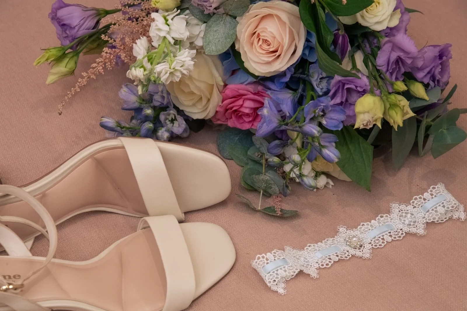 Pair of cream-colored high-heeled shoes, a floral bouquet with pink, purple, and white flowers, and a white lace choker with a pearl embellishment on a pink fabric surface.