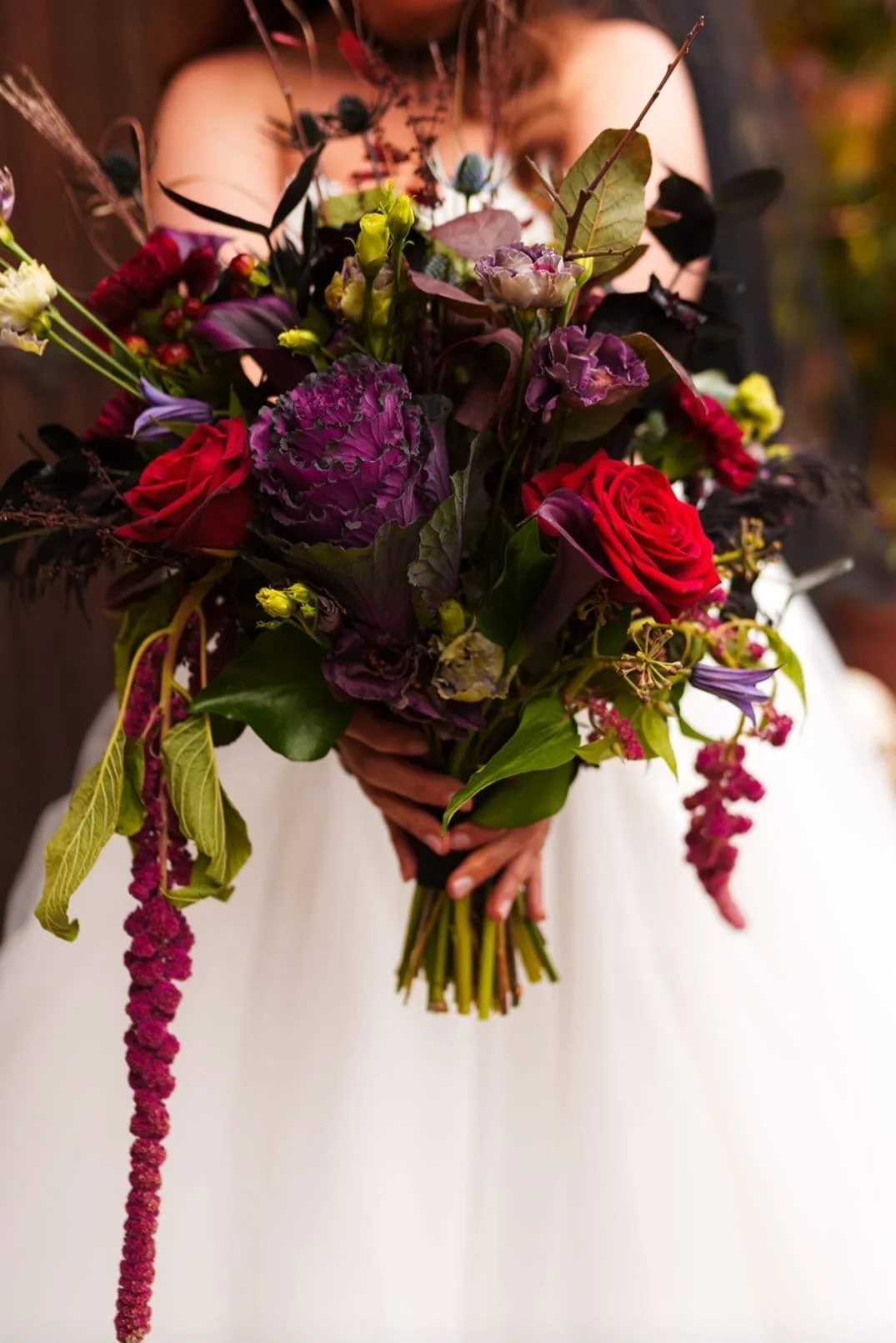 A person holding a large, colorful bouquet of various flowers, including red roses, purple hydrangeas, and other assorted blooms, against a blurred background.