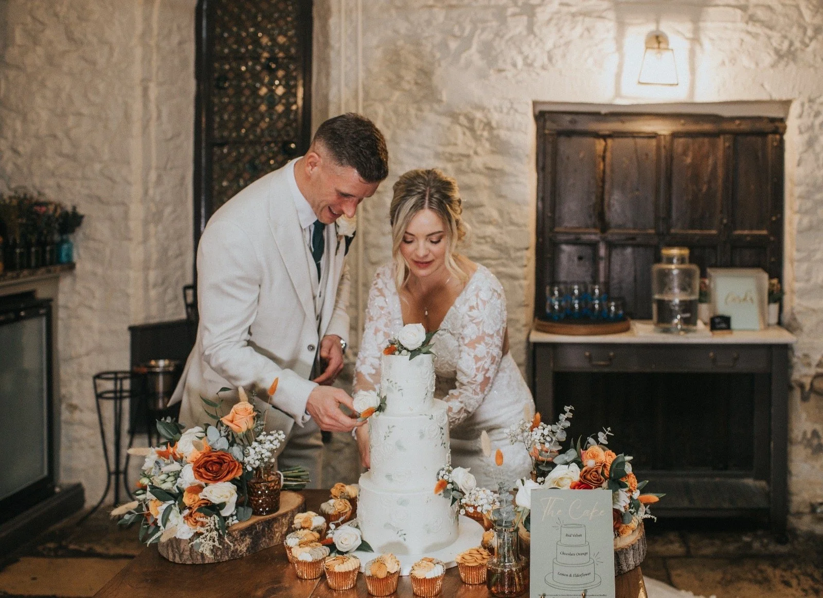 A bride and groom cut a wedding cake together at celebration, with floral arrangements and cupcakes on a rustic table inside a stone-walled room.