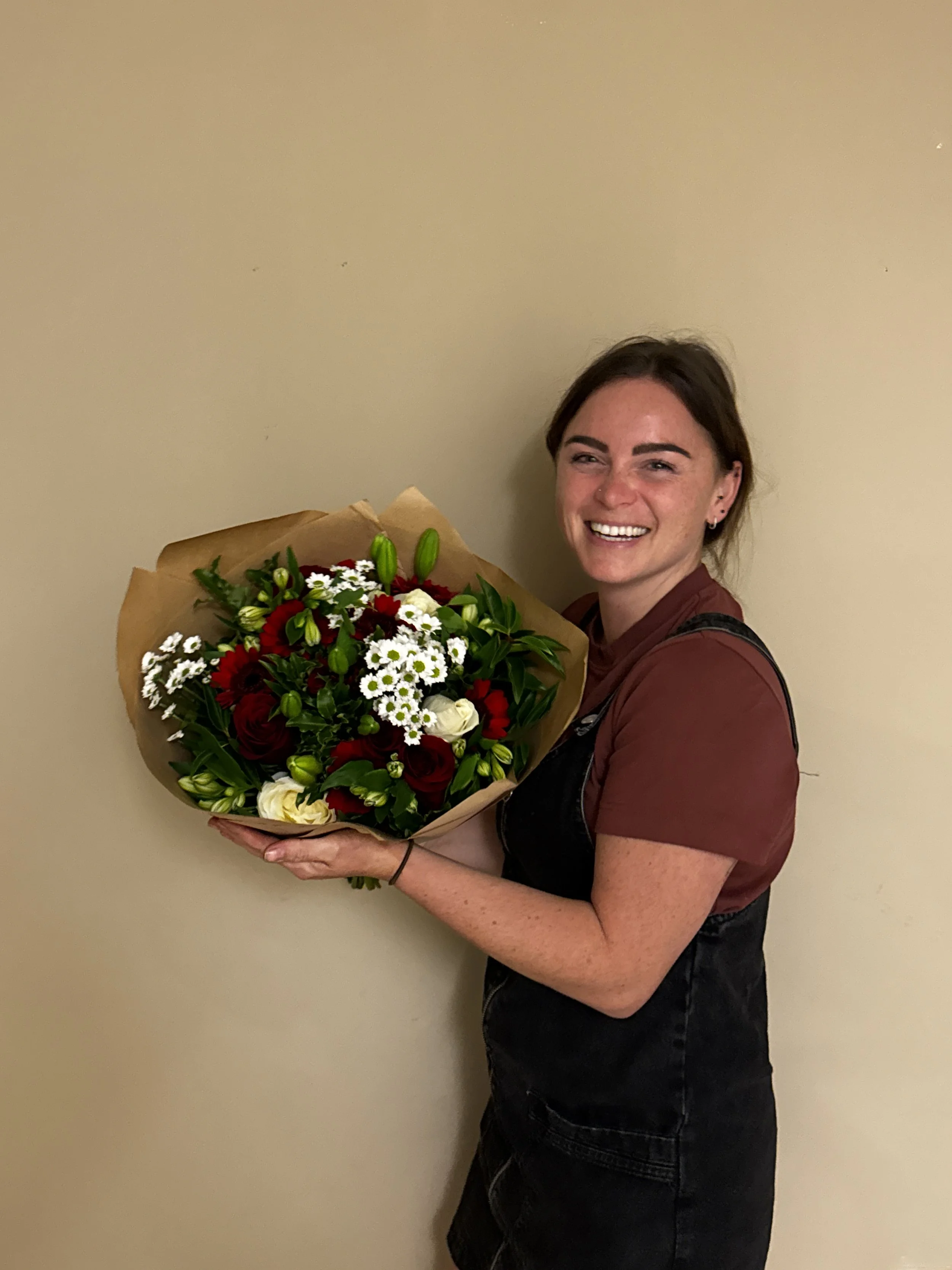 A woman holding a large bouquet of mixed flowers, including white, red, and purple blooms, and smiling at the camera, standing against a plain beige wall.